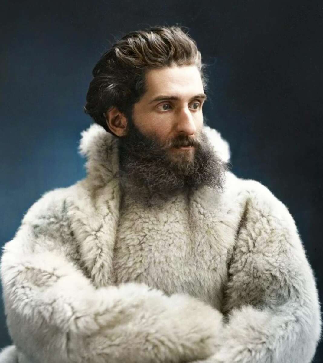 A bearded man with wavy dark hair is wearing a thick, light-colored fur coat, looking slightly to the side against a dark blue background.