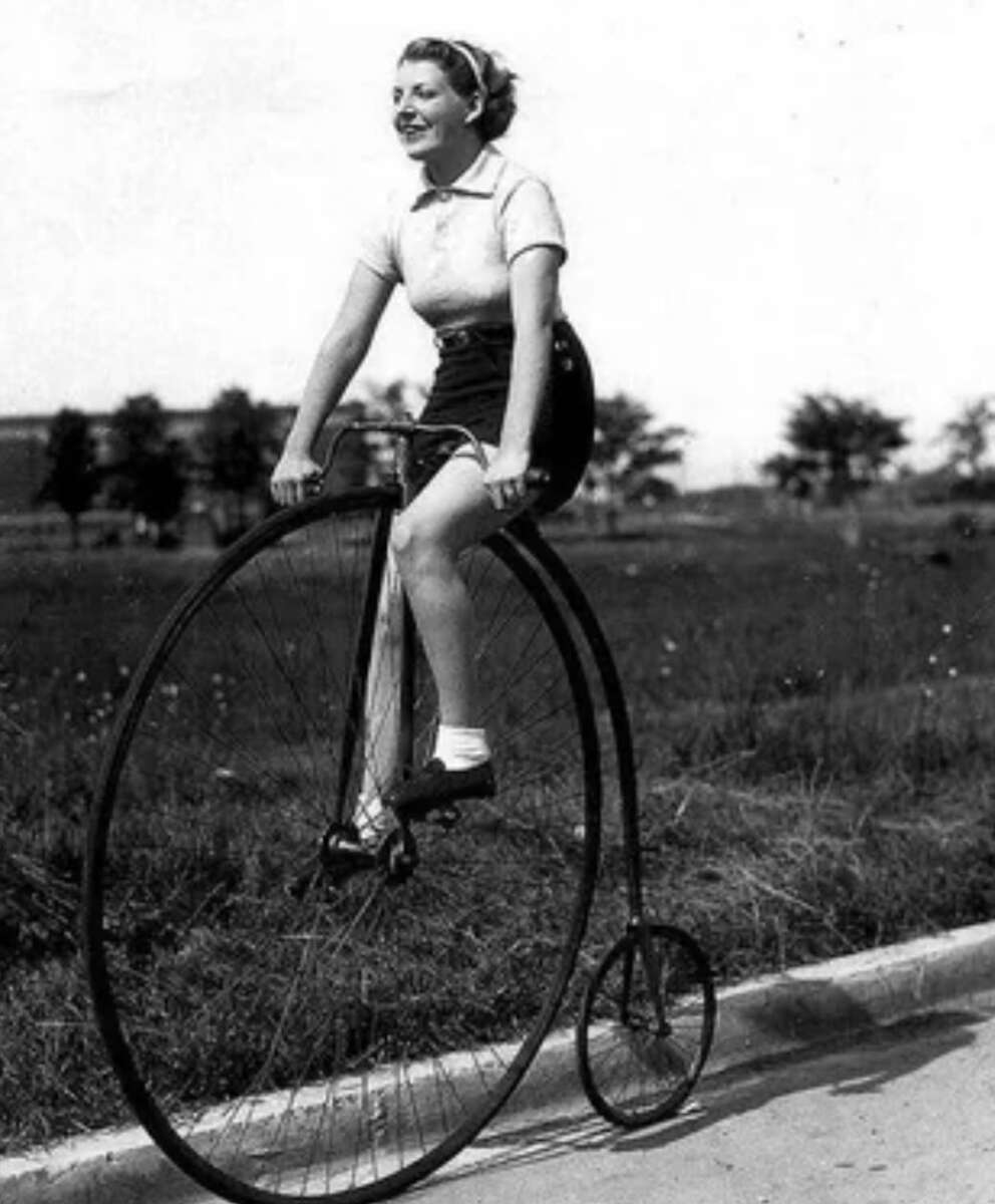 A woman smiles while riding a vintage high-wheeled penny-farthing bicycle on a paved path, with grass and trees in the background.