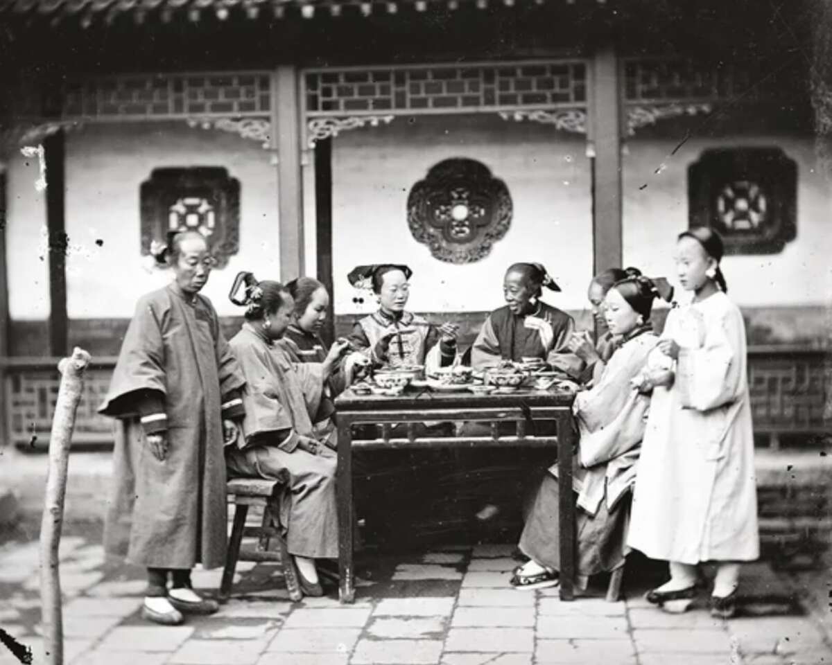 Seven women in traditional Chinese clothing sit and stand around an outdoor table sharing a meal, with ornate windows and a tiled roof visible in the background. The photo appears to be historical, in black and white.