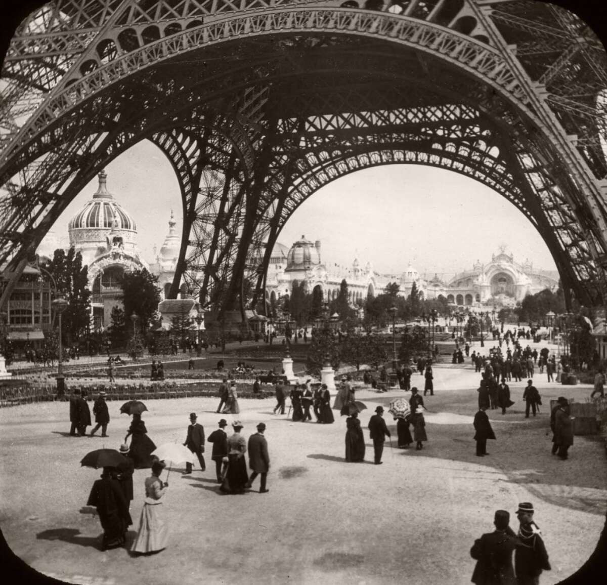 Black-and-white photo of people walking beneath the Eiffel Tower in Paris, wearing early 20th-century clothing, with ornate buildings and trees visible in the background.