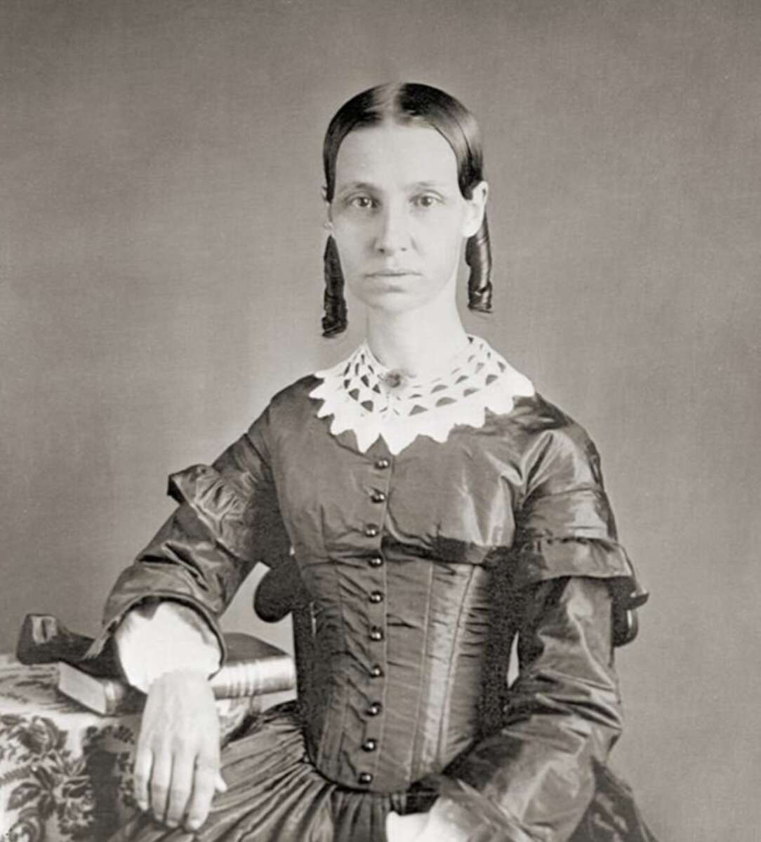 A woman in a dark, long-sleeved 19th-century dress with a white lace collar sits beside a table, one arm resting on a book. Her hair is parted in the center and styled in ringlets. The photo is in black and white.