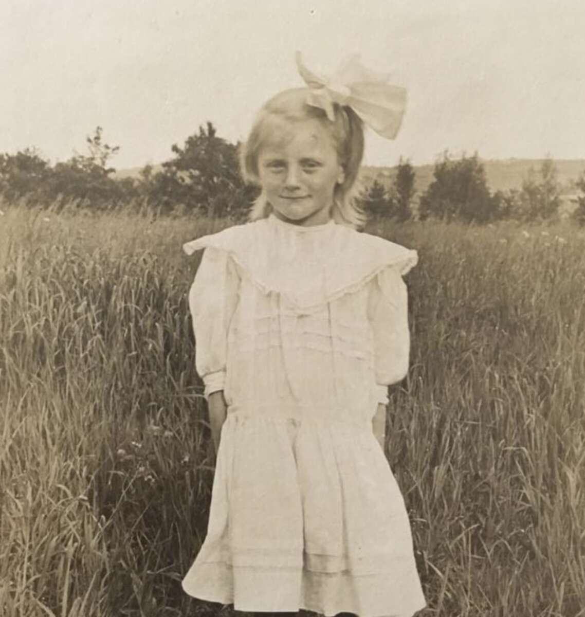 A young girl with light hair, wearing a white dress with a large bow in her hair, stands in a grassy field with trees and sky in the background, smiling softly. The photo is sepia-toned and vintage in appearance.