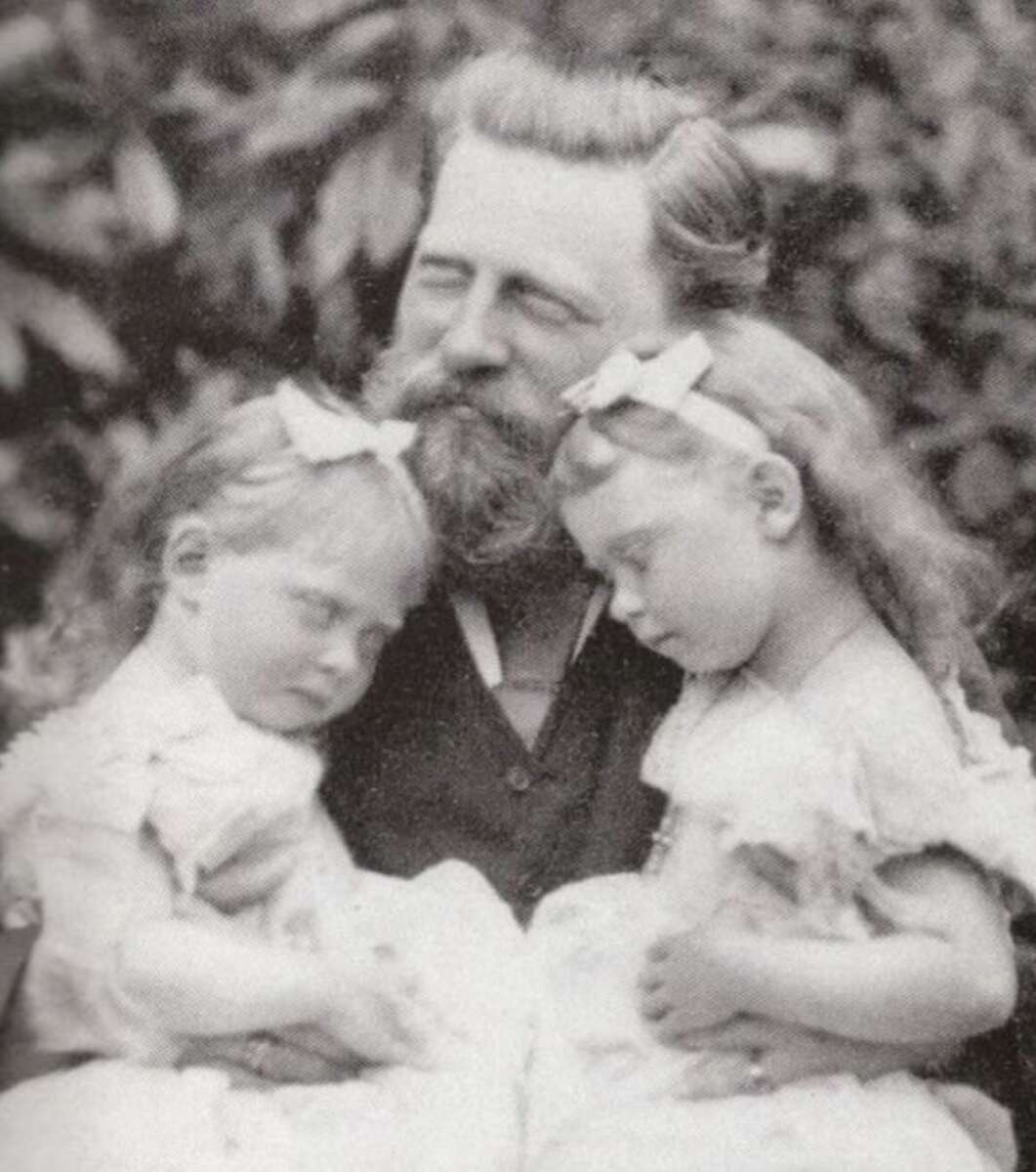A bearded man with closed eyes gently holds two young girls, also with closed eyes and wearing bows in their hair, as they rest against him. The photo is black and white and has a soft, serene atmosphere.