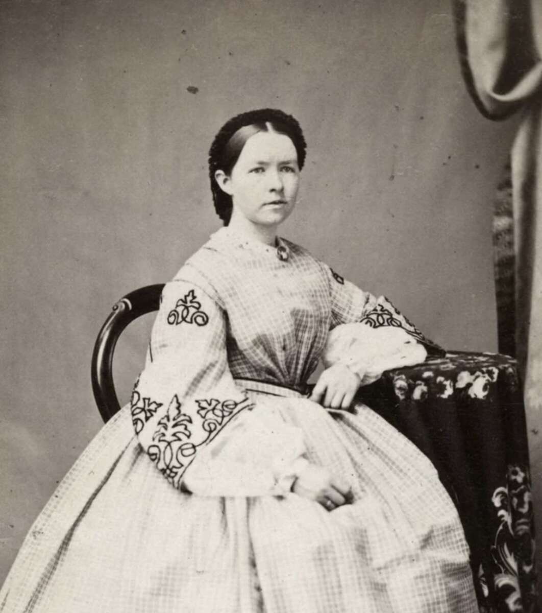 Black-and-white portrait of a seated woman in a 19th-century dress with patterned sleeves, resting one arm on a covered table, looking directly at the camera, with a neutral background.