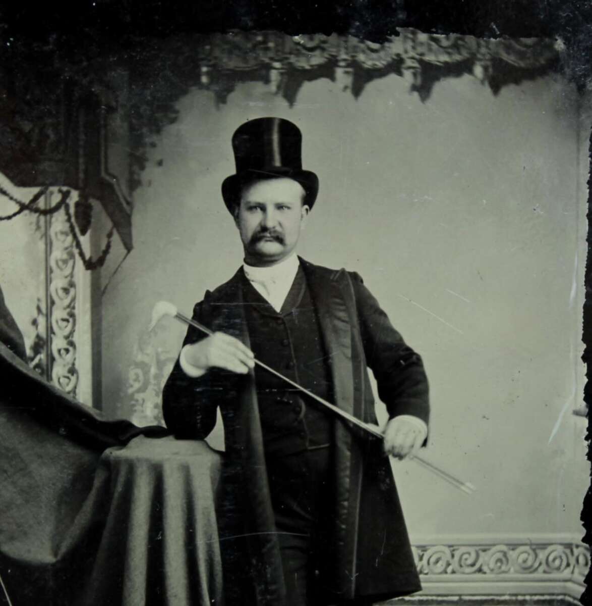 A man with a mustache wearing a top hat and formal suit poses in a vintage studio, holding a cane and resting one arm on a draped table. The backdrop features ornate decorative details.