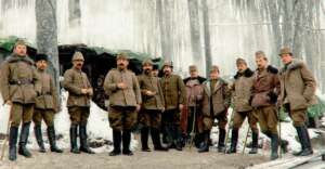 A group of soldiers in winter uniforms and boots stand together in a snowy forest, with icicles hanging from a shelter behind them. The image appears to be historical, from an early 20th-century wartime setting.