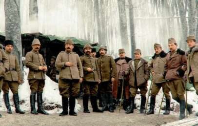 A group of soldiers in winter uniforms and boots stand together in a snowy forest, with icicles hanging from a shelter behind them. The image appears to be historical, from an early 20th-century wartime setting.