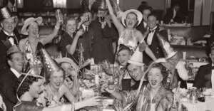 A group of people in party hats and festive attire raise their glasses and smile, celebrating together at a crowded table in a lively, vintage indoor setting.