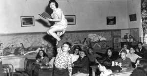 A boy balances a girl on one hand above his head in a classroom, while she holds a book and smiles. Other students sit at desks, watching the performance with interest.