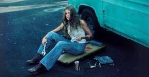 A woman in jeans and boots sits on a mechanic's creeper by a turquoise van, holding a wrench. Tools and a can of soda are on the ground beside her. She appears relaxed, with long hair and a casual expression.