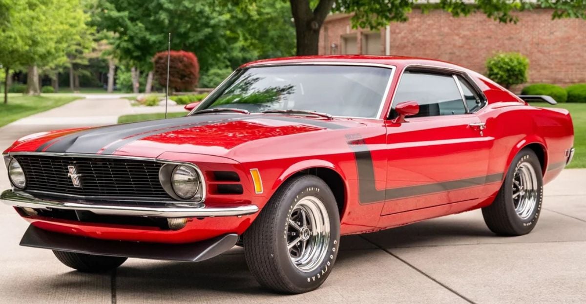 A bright red classic Ford Mustang with black racing stripes is parked on a driveway, surrounded by green trees and manicured lawns in a suburban neighborhood.