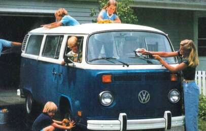 A family washes a blue and white Volkswagen camper van in a driveway. Two kids are on the roof, one adult is painting, two children clean the front, and another washes a tire. A house and white picket fence are in the background.