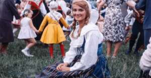 A young woman in traditional dress sits on grass, smiling at the camera. Children and adults, some in costumes, stand and hold hands in a circle in the background at an outdoor event.