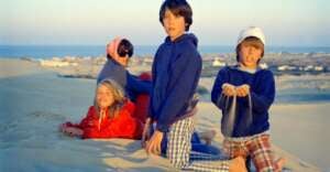 Four children in casual clothes sit and kneel on sandy dunes with a woman in sunglasses. The background shows a coastal landscape with houses in the distance under a clear sky. One child lets sand fall from his hands.