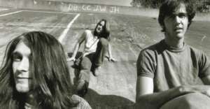 Three young men with long hair sit and relax on a sloped, concrete surface outdoors. The scene is in black and white. Graffiti with initials is visible on the wall behind them.