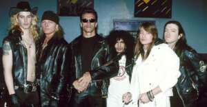 Six men stand together, most wearing black leather jackets. One man in the middle wears sunglasses. Another man wears a white jacket. They pose against a blue wall with colorful artwork in the background.