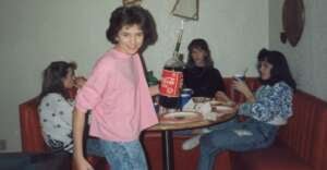 Four teenage girls sit in a red booth around a table with pizza and drinks. One girl in a pink shirt stands smiling, holding a large bottle of Coca-Cola. The others sit, talking or eating.