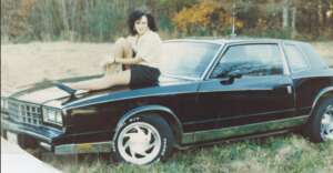 A woman with dark hair sits on the hood of a black classic car parked in a grassy field with autumn trees in the background. She is wearing a light top and a dark skirt, and is looking at the camera.