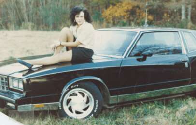 A woman with dark hair sits on the hood of a black classic car parked in a grassy field with autumn trees in the background. She is wearing a light top and a dark skirt, and is looking at the camera.