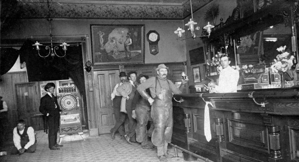 Black-and-white photo of men in work clothes gathered at a bar in an ornate room with a bartender. Decorative art, a clock, and a chandelier are visible; one man sits on the floor at left near a large safe.