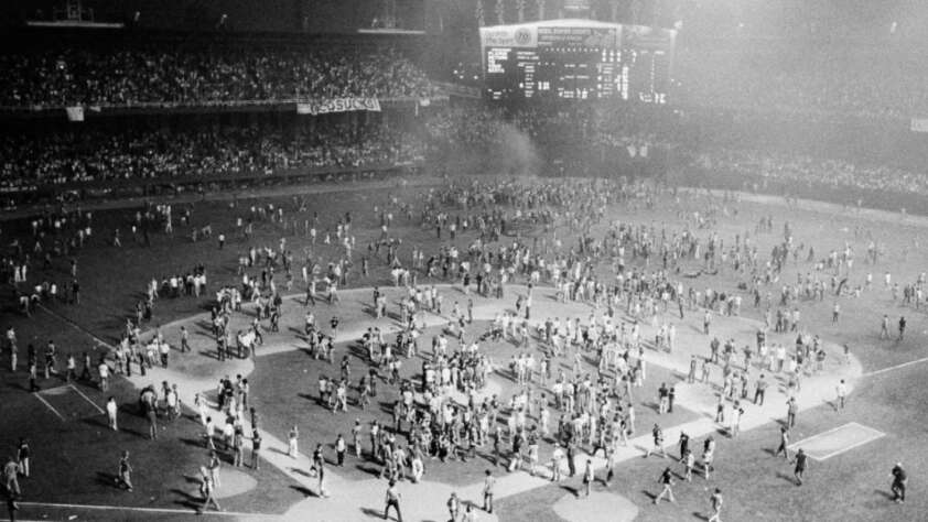A large crowd of people rush onto a baseball field at night, filling the infield and outfield areas, while stadium lights illuminate the scene and spectators watch from the stands.