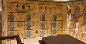 Ancient Egyptian tomb walls decorated with colorful hieroglyphs, gods, and twelve blue baboon figures. An ornate sarcophagus sits in the foreground, partially visible. Warm lighting highlights the detailed artwork.