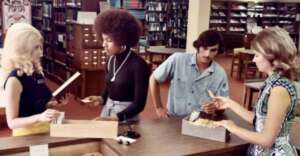 Four people stand at a library counter, using card catalogs to search for books. Bookshelves fill the background, and the setting appears to be from the 1970s based on their clothing and hairstyles.