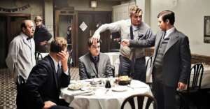 Five men in suits are in a tense scene at a restaurant; one man angrily grabs another&rsquo;s head while a third points a gun at him. Two other men watch, one seated with his hand on his face. Food and drinks are on the table.