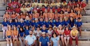 A large group of children and adults wearing matching blue, orange, red, and yellow t-shirts sit on outdoor bleachers, posing and smiling for a group photo. The setting appears to be a summer camp or school event.