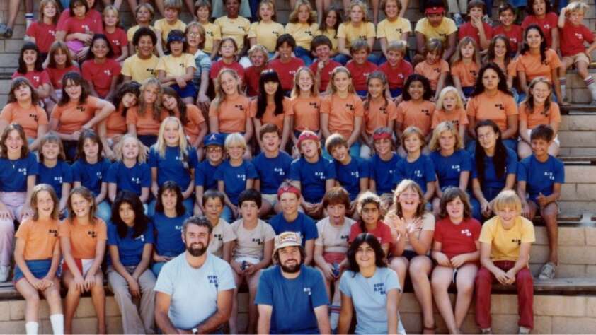 A large group of children and adults wearing matching blue, orange, red, and yellow t-shirts sit on outdoor bleachers, posing and smiling for a group photo. The setting appears to be a summer camp or school event.