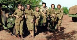 A group of seven women in military uniforms pose together outdoors, smiling beside and sitting on a military jeep. Trees and another army vehicle are visible in the background.