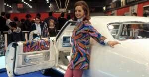 A woman in a colorful patterned coat and pink tights poses beside a white classic car with matching patterned interior at an indoor car show. People and exhibition signs are visible in the background.