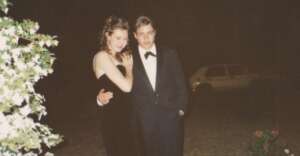 Two young people dressed formally, a woman in a black dress and a man in a black tuxedo, stand outside at night. The man has his arm around the woman. There are white flowers and a car in the background.