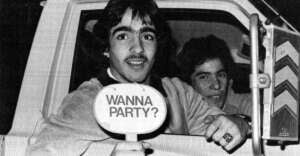 Two young men sit in a car at night. The man in front holds a sign outside the window that reads "WANNA PARTY?" Both appear to be smiling or laughing, creating a fun and lively atmosphere.