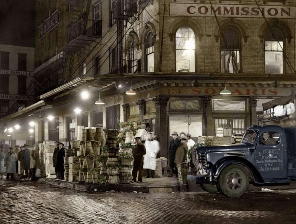 A vintage street scene shows workers stacking baskets of produce outside a market at night. A blue delivery truck is parked nearby, and people stand along the sidewalk by a brick building with "COMMISSION" and "SOLARIS" signage.