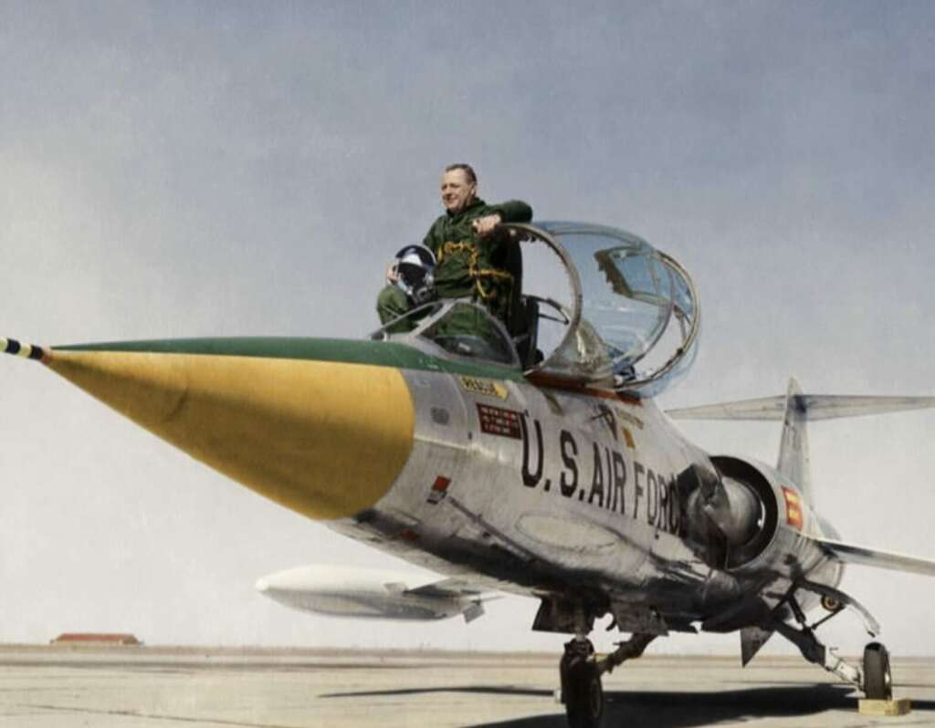 A pilot and a crew member sit in the open cockpit of a U.S. Air Force jet with a yellow-tipped nose, parked on an airfield under a clear sky.