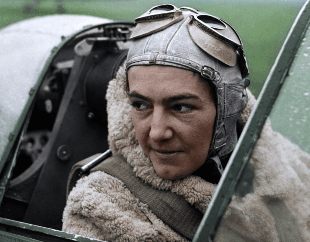 A pilot in a leather flying helmet and goggles sits in the cockpit of an aircraft, wearing a thick shearling flight jacket and looking determinedly to the side.