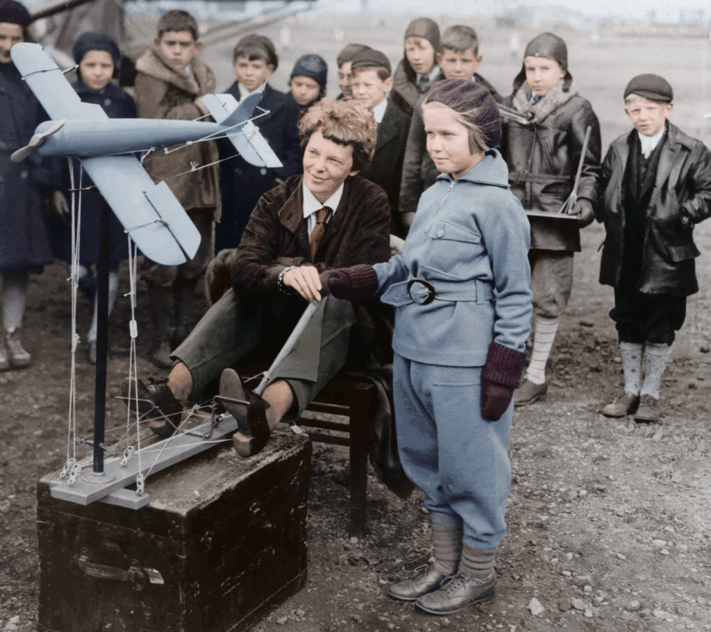 A woman and a young girl operate a small model airplane outdoors, surrounded by a group of children dressed in early 20th-century clothing, with some watching attentively in the background.