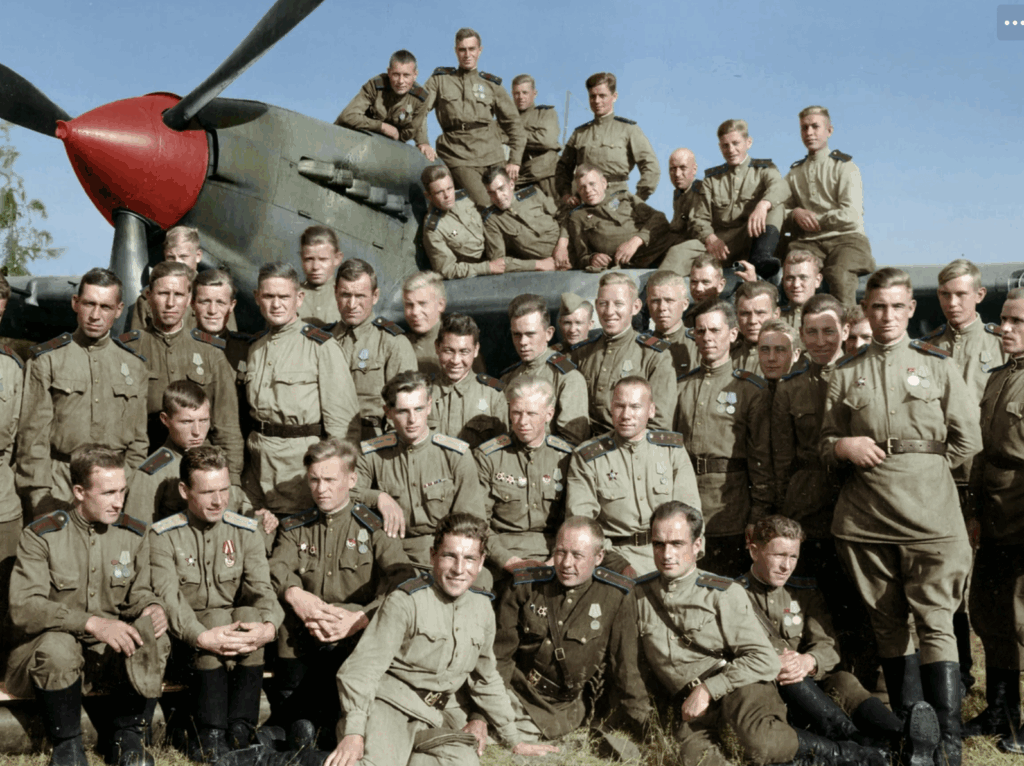 A large group of uniformed soldiers pose for a photo in front of a military aircraft with a red nose, outdoors on a sunny day. Some men sit on the plane, while others stand or crouch on the grass.
