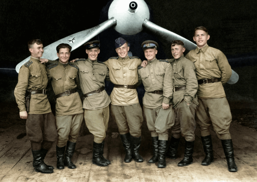 A group of seven young men in vintage military uniforms stand smiling with arms around each other in front of a large airplane propeller.