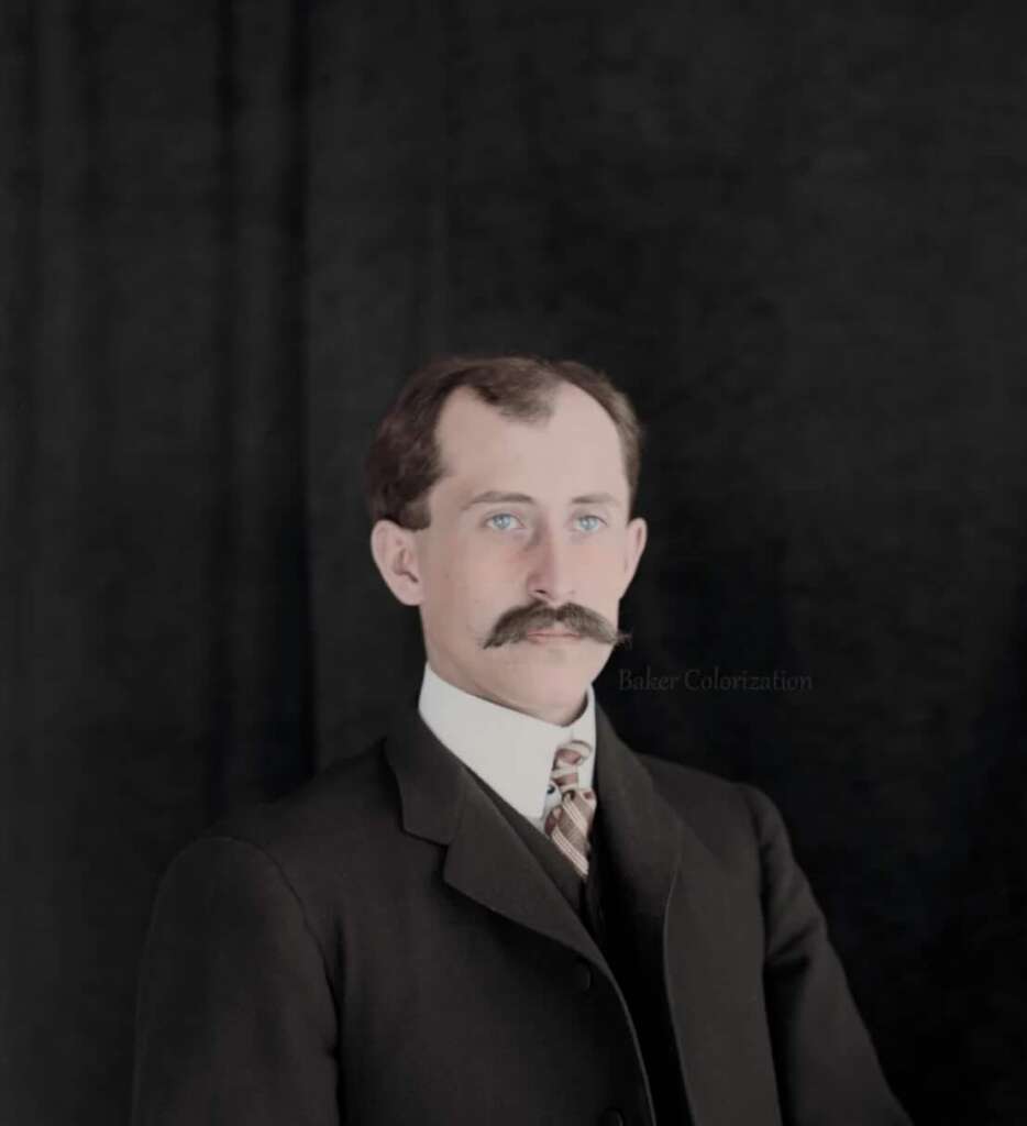A man with light skin, blue eyes, and a dark mustache wears a black suit, white shirt, and patterned tie. He poses against a plain black background. The image appears to be an old portrait that has been colorized.