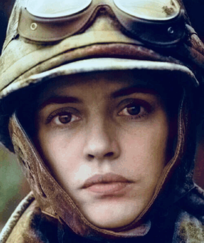 A person with a serious expression wears vintage aviator goggles and a helmet, with a close-up focus on their face. The background is softly blurred, highlighting the facial details and headgear.