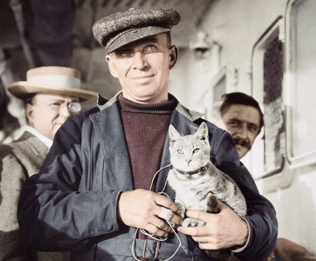 A man in a cap and coat smiles while holding a gray cat. Two other men stand in the background, one wearing a hat, on what appears to be a ship or boat.