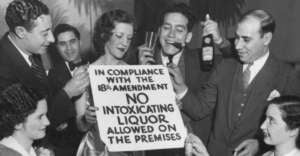 A group of smiling people at a party hold drinks and cigarettes, surrounding a woman holding a sign that reads, "In compliance with the 18th Amendment, no intoxicating liquor allowed on the premises.
