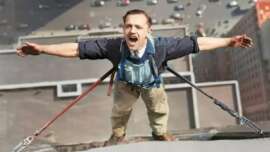 A man stands on the edge of a tall building, secured by safety harnesses, with his arms outstretched and mouth open, possibly shouting or laughing, overlooking a city street far below.