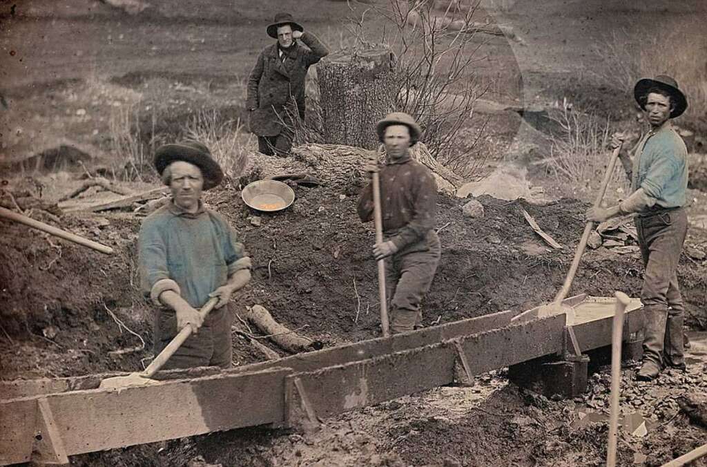 Four men work outdoors at a mining site, using tools to sift through soil in a wooden sluice box. They wear old-fashioned clothing and hats; one man stands in the background observing the scene.