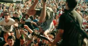 A person does a backflip amid a lively crowd at an outdoor concert, with people cheering and a musician performing on stage in the foreground.