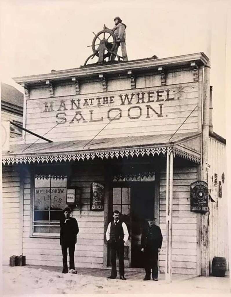 Black-and-white photo of three men standing in front of a wooden saloon called “Man at the Wheel Saloon,” with a man on the roof holding a large ship’s wheel. Signs for beer are visible in the windows.