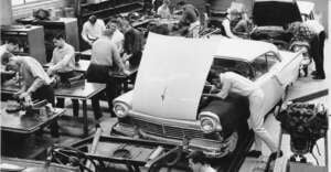 Black and white photo of students working on car engines and parts in an auto shop classroom, with one group focused on a car with its hood up and others working at tables with various tools and engine components.