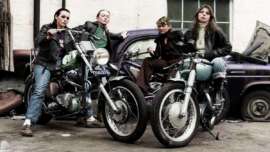 Four women in leather jackets pose confidently with vintage motorcycles in front of an old car and a brick wall. The scene has a rebellious, retro vibe.